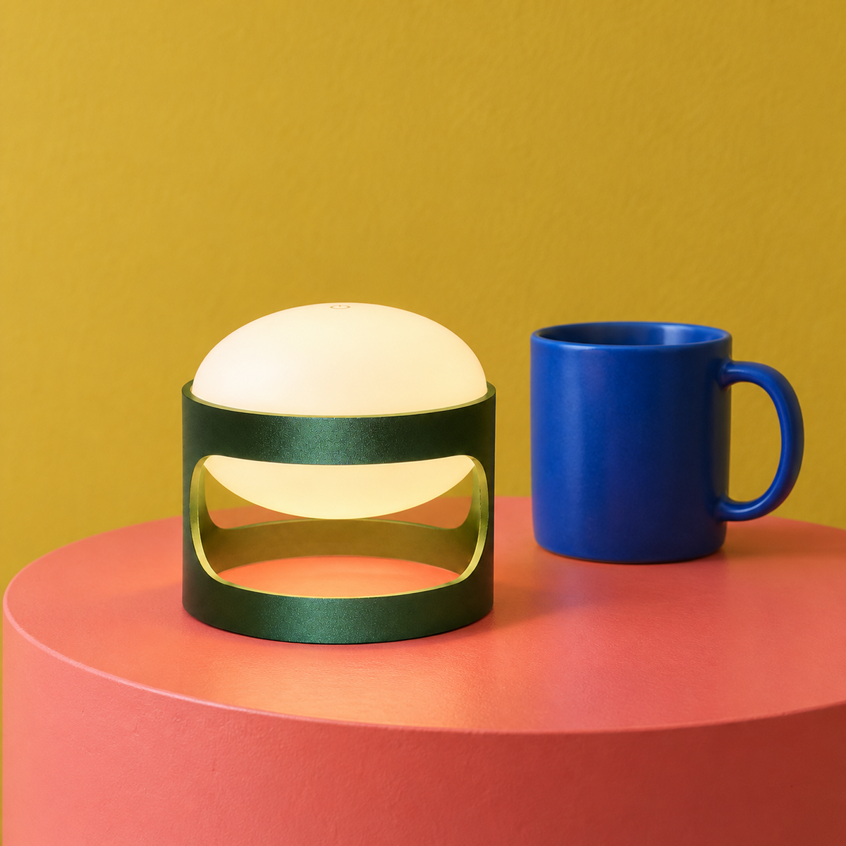 Lotus Mini cordless lamp in Deep Forest Green on a coral plinth beside a blue mug for scale