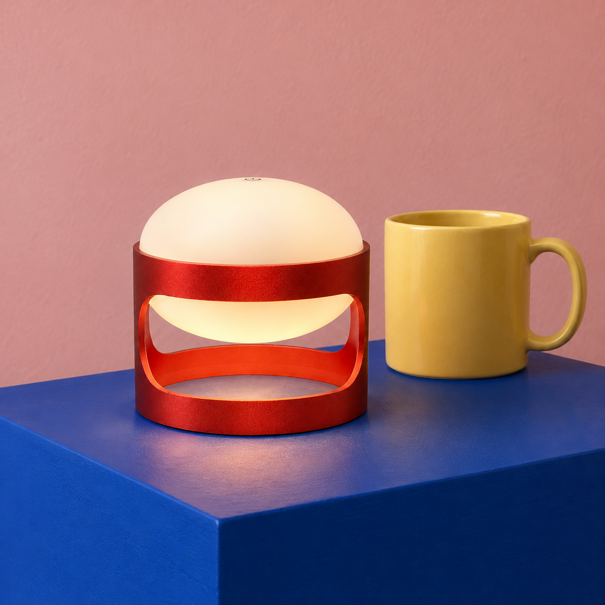 Lotus Mini cordless lamp in Deep Red on a cobalt blue block beside a yellow mug for scale